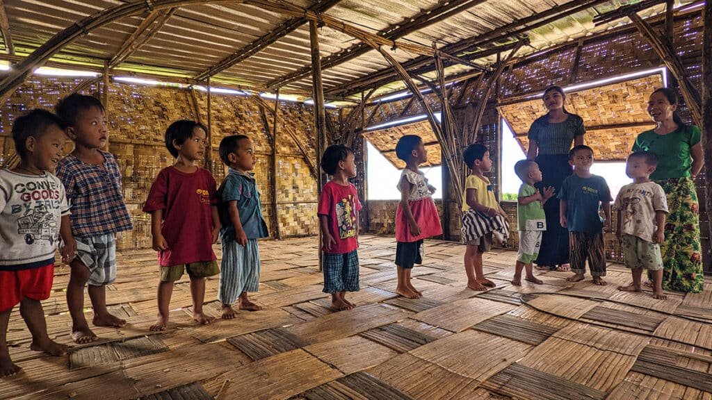 Inside a bamboo home in Myanmar, children stand barefoot on woven floors while two women smile nearby — a warm, everyday moment inside a resilient, community built shelter.