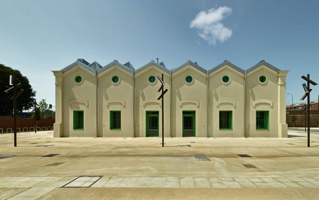 The restored facade of a historic building in Milan&rsquo;s Olympic Village, featuring cream-colored walls and vibrant green windows under a bright blue sky.
