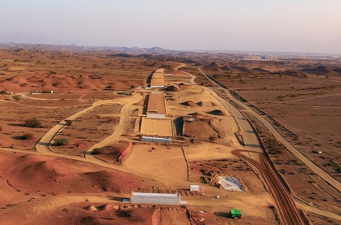 Aerial view of a massive desert railway construction site, with long stretches of graded earth and temporary structures winding through red sand dunes under a hazy sky.