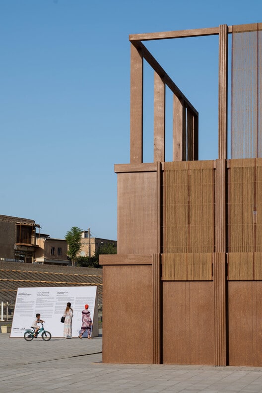 The AlMusalla Pavilion&rsquo;s wooden frame stands tall under a clear blue sky in Bukhara, its woven panels catching the sun. In the background, locals stroll by &mdash; a child on a bike, women walking &mdash; turning the architectural installation into part of daily life.
