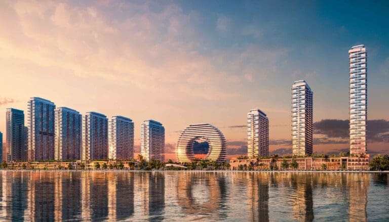 مشهد غروب ساحر لأبراج داون تاون أم القيوين، حيث تتلألأ الأبراج الحديثة تحت ألوان السماء الوردية والذهبية، وتنعكس صورتها على الماء الهادئ، مع برج دائري فريد يُشكّل نقطة تركيز في المشهد.