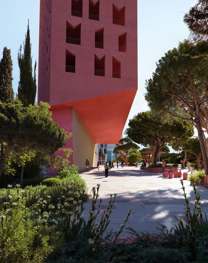 A low-angle view of the bold red Vlore Beach Tower, showcasing its sculptural base and recessed openings, framed by lush green trees, white flowers, and people strolling through the sun-dappled plaza below.