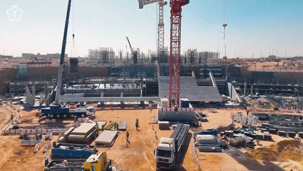 صورة جوية لمشروع بناء ضخم تحت الشمس الساطعة، تظهر فيها رافعات عملاقة، عمال يرتدون سترات أمان، ومدرجات خرسانية قيد الإنشاء، مع شاحنات وأكوام من مواد البناء في موقع العمل.