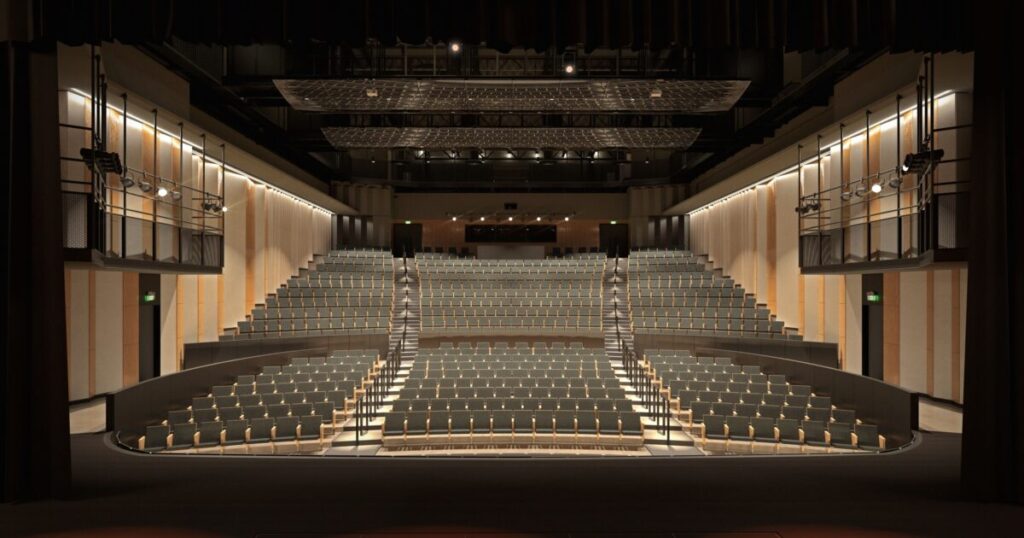 A modern, minimalist auditorium with tiered gray seats, warm wood paneling, and sleek lighting fixtures. The space feels open, functional, and designed for contemporary performances.