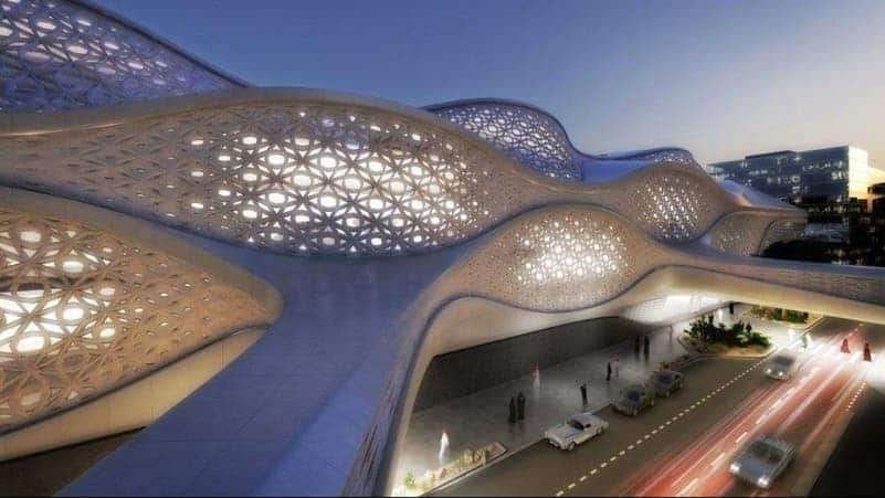 An evening view of the King Abdullah Financial District Metro Station, showcasing its glowing, patterned concrete canopy that resembles traditional Islamic latticework. Below, cars streak by in light trails while pedestrians walk under the illuminated structure.