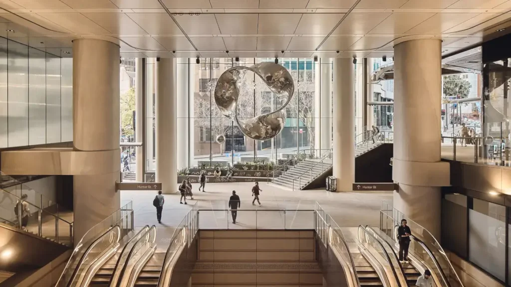 The airy lobby of 1 Elizabeth Tower, with sweeping escalators, giant sculptural art hanging from the ceiling, and people moving through a sunlit, column-framed space.