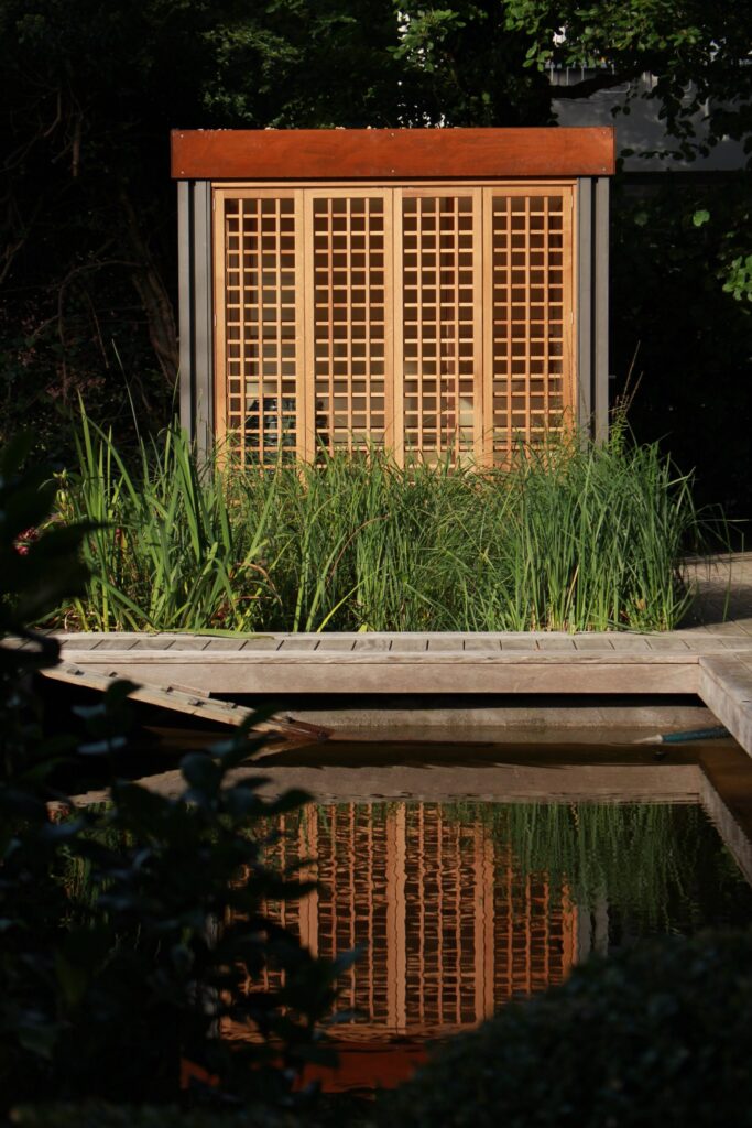 Wooden sauna with lattice doors, reflected in pond water, surrounded by tall grasses.

Caption: