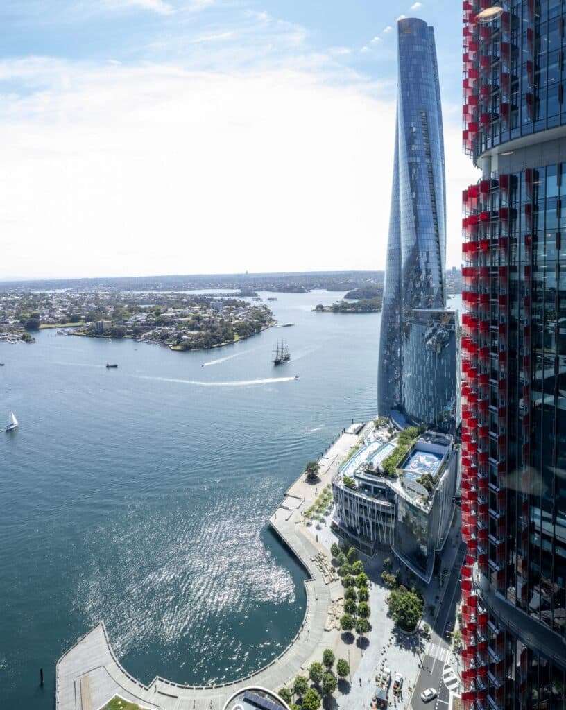 Aerial view of Barangaroo: glass towers overlook the harbour, with boats gliding across sunlit water.