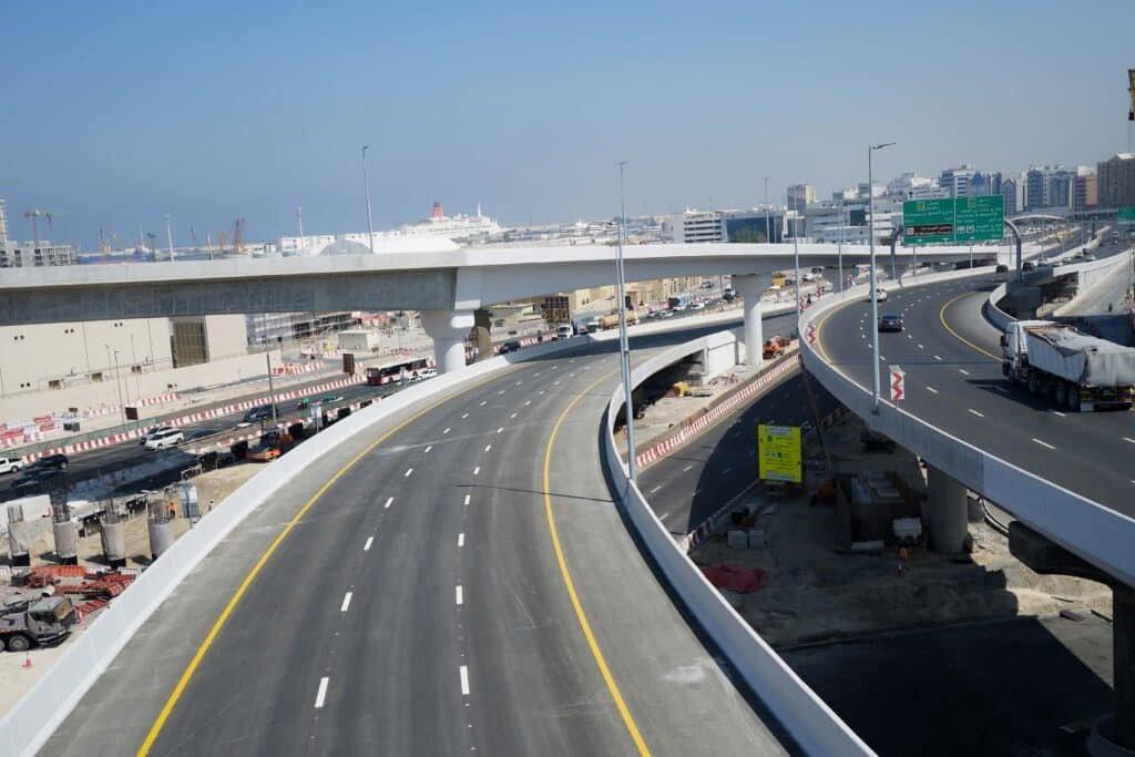 A sleek new overpass on a major highway in Dubai, curving gracefully above traffic with clear signage, construction zones nearby, and a backdrop of port cranes and massive cruise ships under a bright blue sky.