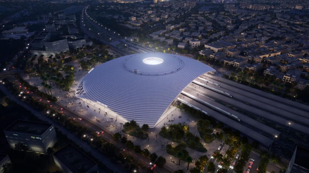 A nighttime aerial view of Casablanca&rsquo;s new Casa Sud Station, showcasing its glowing white dome, sleek trains on adjacent tracks, and crowds of people strolling around the landscaped plazas and tree-lined walkways.