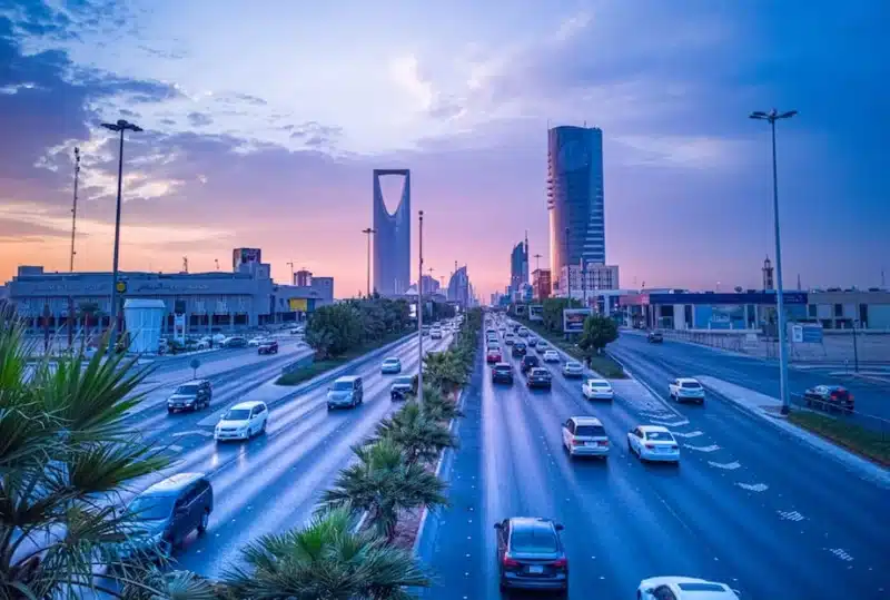 A stunning urban sunset view of Riyadh, featuring iconic towers like Kingdom Tower and Faisaliah Tower, with busy highways lined by palm trees &mdash; capturing the city&rsquo;s dynamic growth and modern energy.