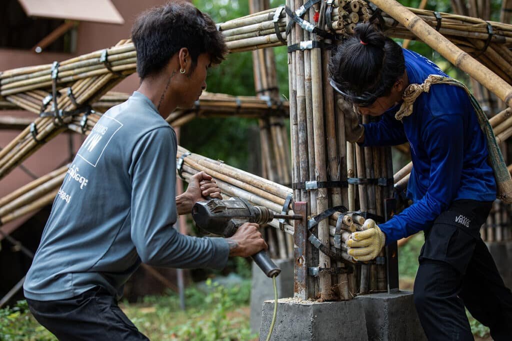 Two builders in Myanmar work together to assemble a bamboo frame — one drilling, the other steadying the structure — showing teamwork and local skill in creating earthquake-resistant homes.