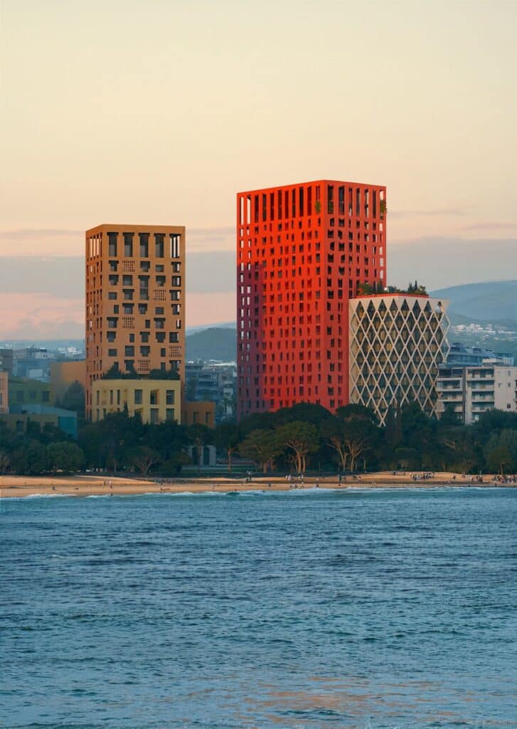 The vibrant red Vlore Beach Tower glows at sunset, standing tall beside other architecturally distinct buildings, with a sandy beach full of people in the foreground and calm sea waters reflecting the golden sky.
