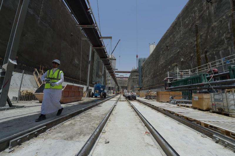 A worker in traditional Saudi attire and a high-visibility vest walks alongside newly laid railway tracks inside a tunnel or station under construction, with cranes and construction equipment visible in the background.