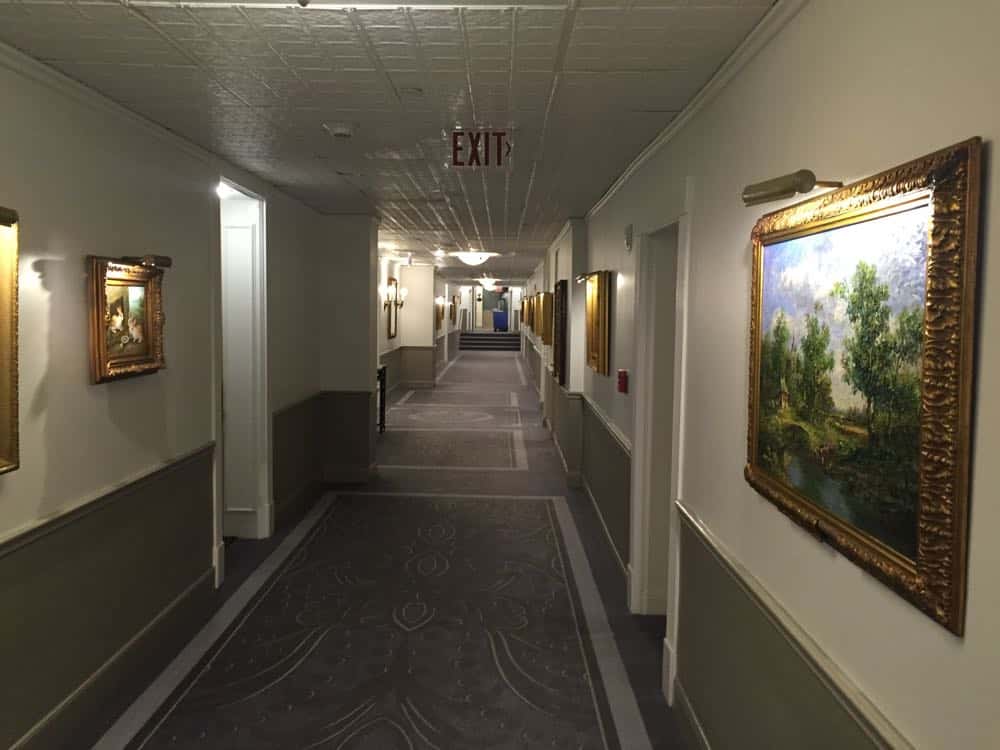 A long hotel corridor at The Driskill, with patterned carpet, white walls adorned with gold-framed paintings, and an &ldquo;EXIT&rdquo; sign glowing overhead.
