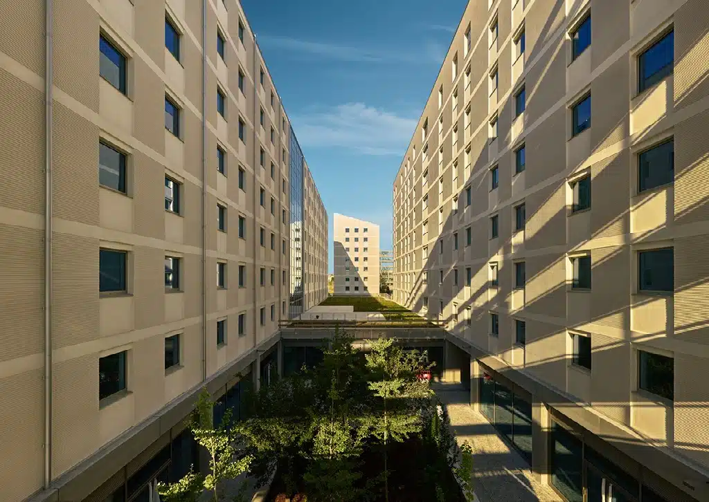 A narrow courtyard between two modern residential buildings in Milan&rsquo;s Olympic Village, with small trees growing in the center and sunlight casting long shadows on the walls under a clear blue sky