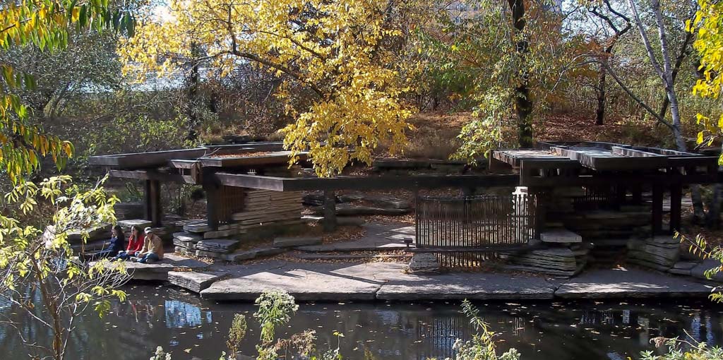 Three people sit quietly on stone steps beside a pond under golden autumn trees, framed by wooden pavilions and fallen leaves.
