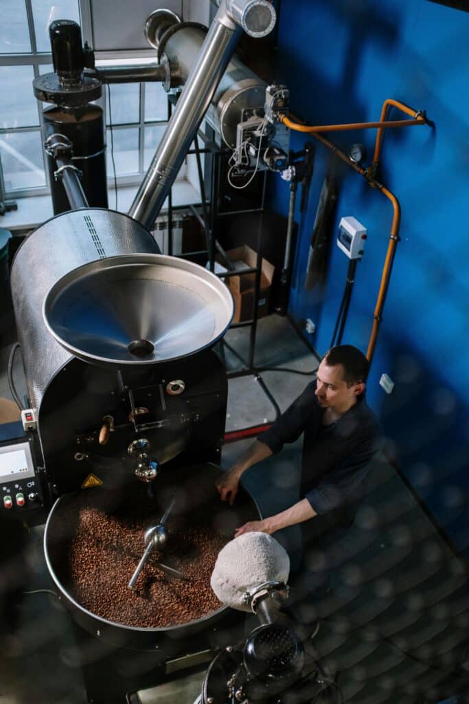 A man watches freshly roasted coffee beans cool in a modern roaster &mdash; the quiet moment where waste begins its second life as a sustainable building material.