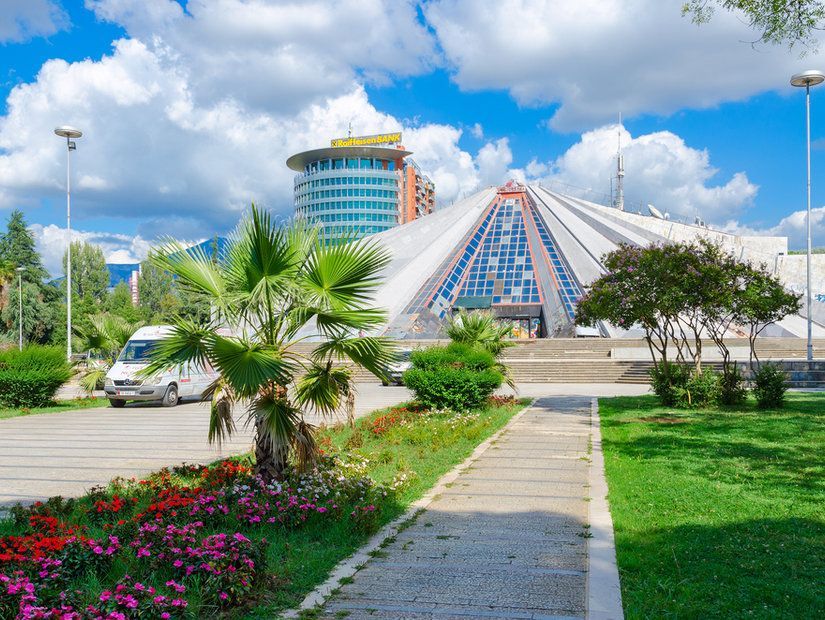 A striking geometric building in Tirana with a glass pyramid facade, surrounded by greenery and a paved walkway under blue skies.