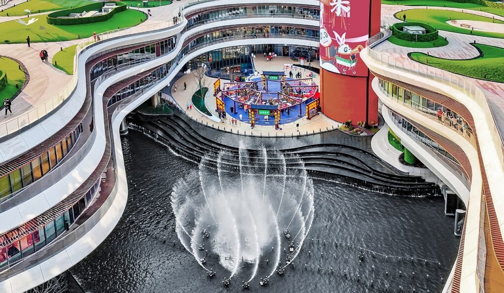 Aerial view of Chengnan’s river heart: fountains dancing, curved walkways winding, green rooftops above, and people enjoying the plaza below.