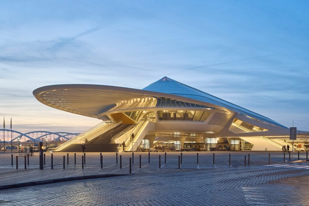 Mons station exterior at dusk — futuristic, glowing white structure under a soft blue sky.