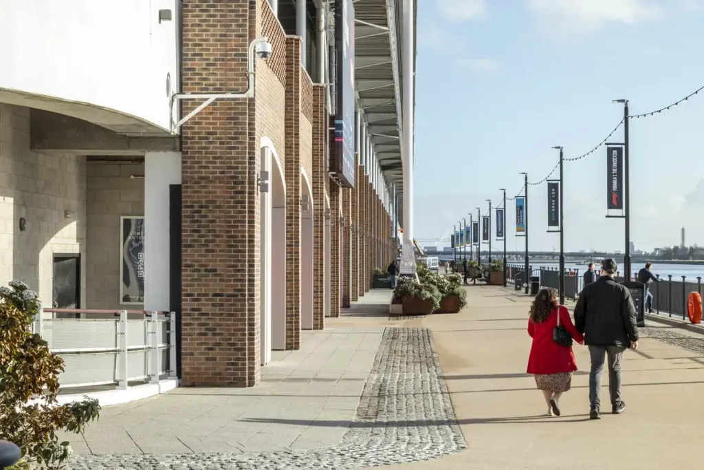 A couple strolls hand-in-hand along the riverside walkway of Immerse LDN, passing under brick arches and modern banners. The sun casts long shadows on the pavement, while the Thames glimmers in the distance &mdash; a quiet moment in a space built for connection.