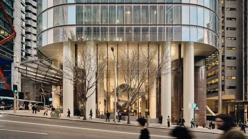 The grand entrance of 1 Elizabeth Tower, framed by towering white columns and glass walls, with bare trees and blurred pedestrians adding life to the urban streetscape.