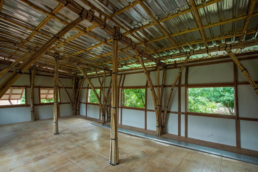 Inside a bamboo-framed structure in Myanmar: tall, slender poles support a corrugated roof, with large windows framing green trees outside simple, airy, and built to last.