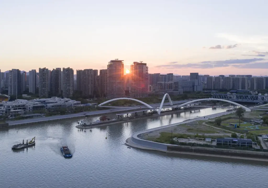 Sunset over Hangzhou’s Grand Canal Gateway Bridge — the white structure glows between water and skyline, as boats glide beneath in quiet rhythm.