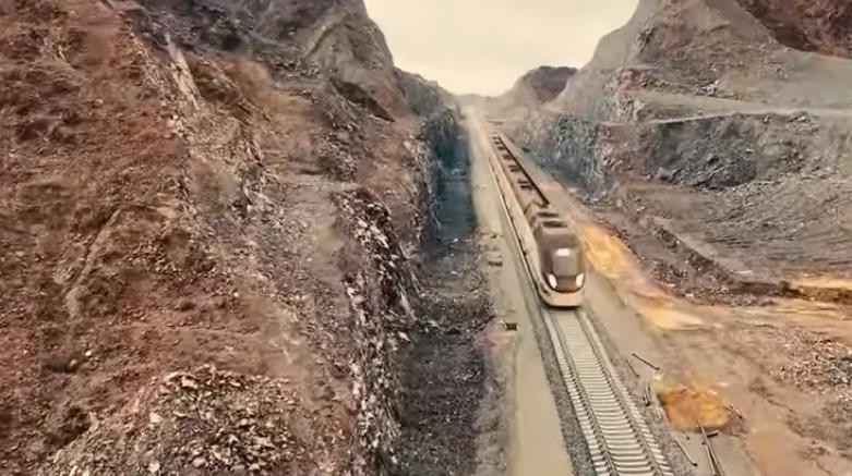 A sleek modern train glides through a narrow mountain valley, flanked by rugged rocky cliffs under a soft overcast sky.