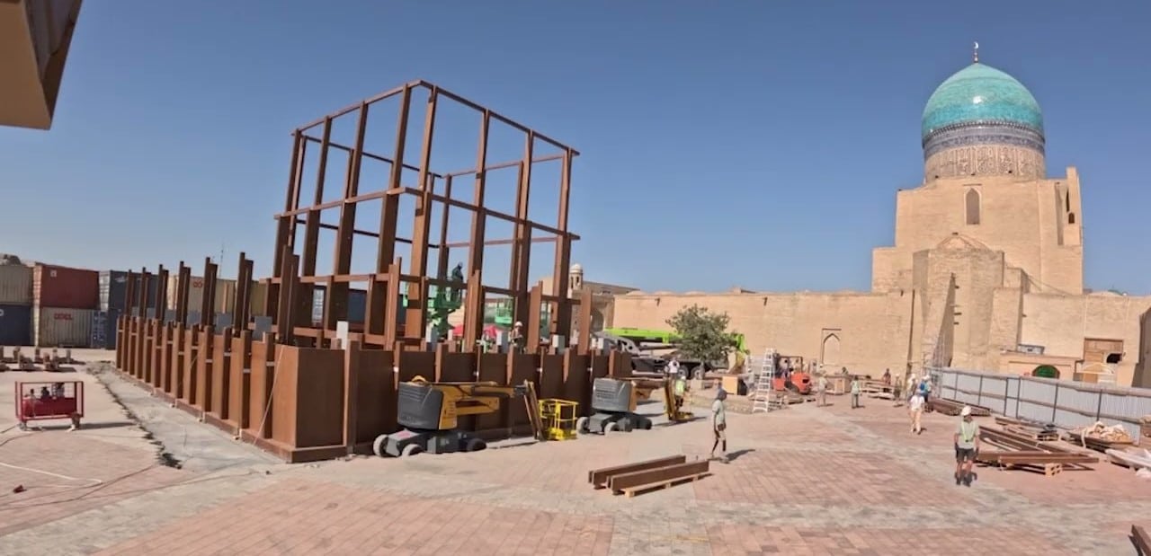 The skeletal frame of the AlMusalla Pavilion rises under a clear blue sky in Bukhara, with workers assembling its modular structure. In the background, the iconic turquoise dome of a historic mosque stands as a silent witness to this modern architectural journey.