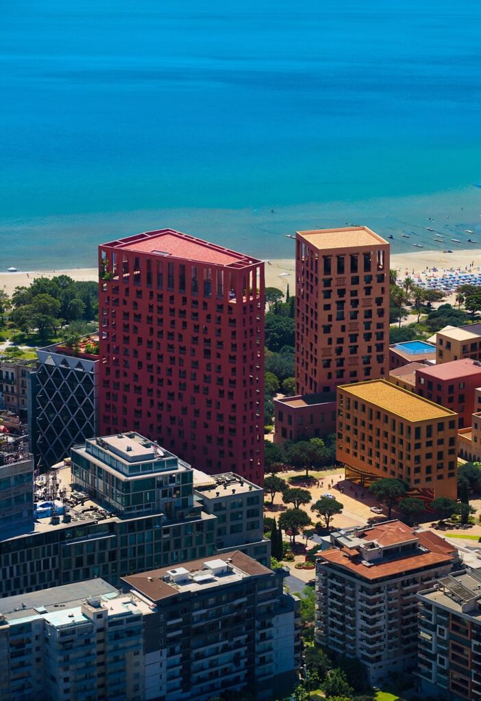 An aerial view of the bold red Vlore Beach Tower rising above the coastal cityscape, surrounded by modern buildings and lush greenery, with a sandy beach and turquoise sea stretching into the horizon under bright sunlight.