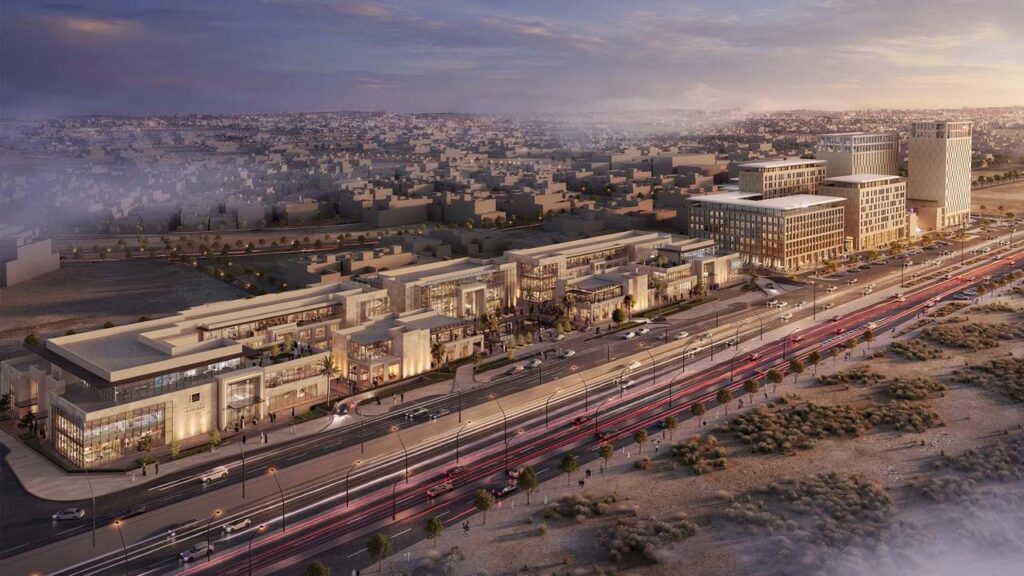 A wide aerial view of a mixed-use development in North Riyadh at dusk &mdash; featuring modern commercial and office buildings, a busy main road with light trails from moving cars, and the sprawling cityscape fading into the horizon.