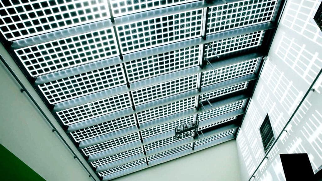 Looking up at a modern building&rsquo;s ceiling with grid-patterned semi-transparent panels that let daylight filter through, casting geometric shadows on the walls.