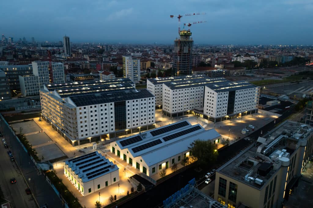 An aerial twilight view of Milan&rsquo;s Olympic Athletes&rsquo; Village, showing modern white buildings with solar panels glowing warmly alongside restored historic structures, set against the city skyline and dusky sky.