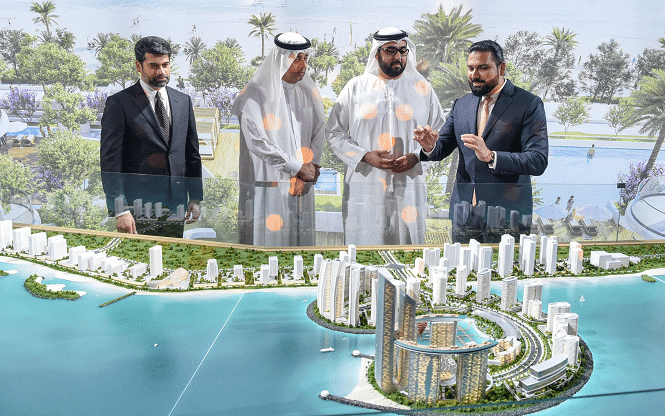 Four men &mdash; two in formal suits, two in traditional Emirati attire &mdash; stand around a large architectural model of Downtown Umm Al Quwain, discussing its design. One gestures as he explains, while the others listen intently. Behind them, a backdrop displays the full waterfront vision of towers, islands, and palm-lined shores.