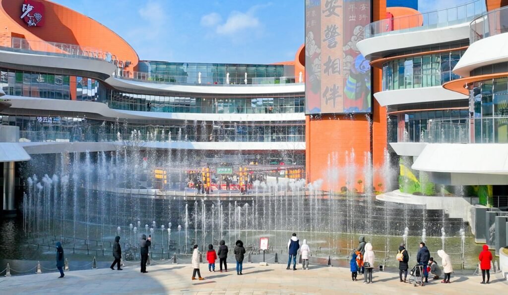 People gather before a grand fountain at Chengnan’s plaza, framed by curved orange buildings and clear blue skies.