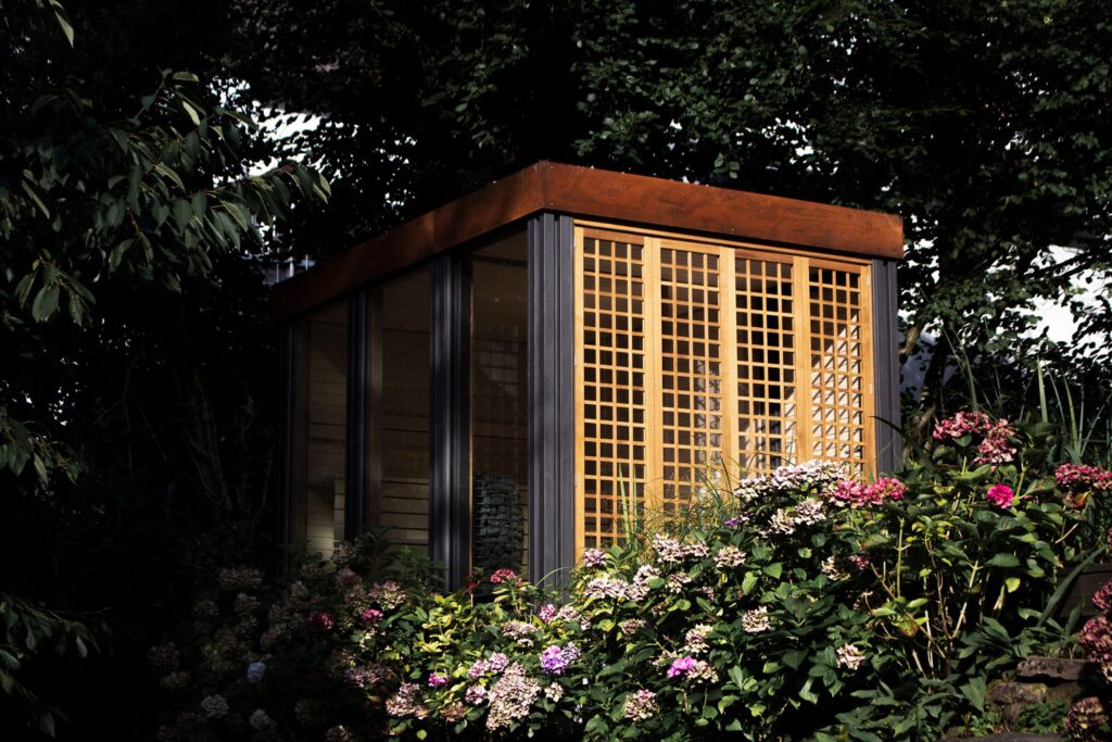 Wooden sauna nestled among hydrangeas and trees, lattice windows glowing softly.