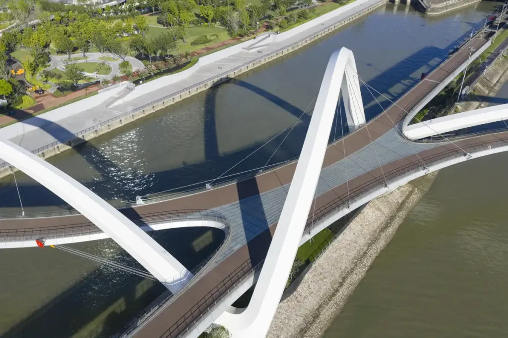Aerial view of the Grand Canal Gateway Bridge — interwoven white arches casting reflections on water, flanked by green parks and urban pathways.
