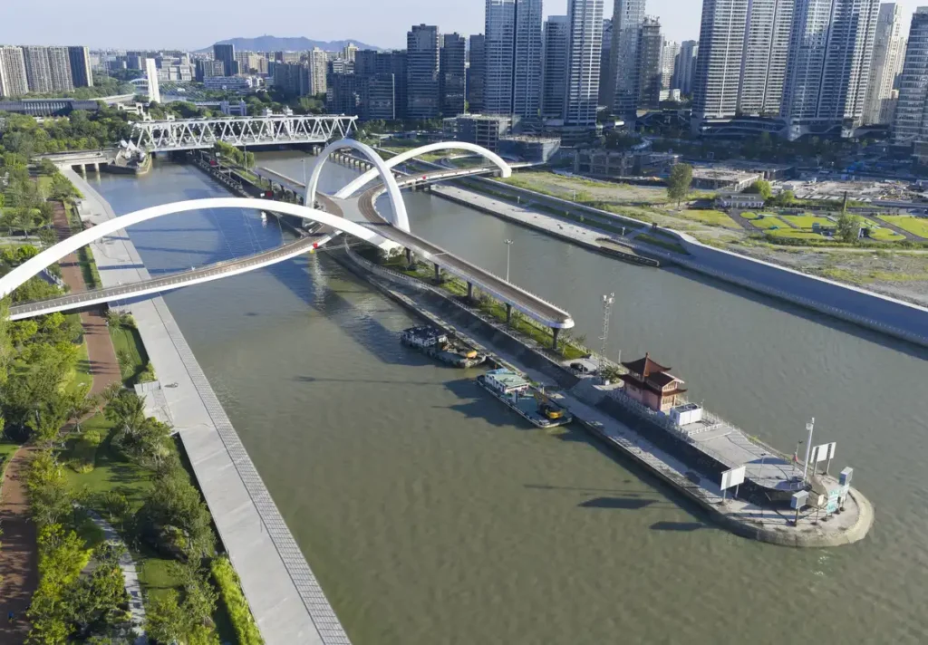 Aerial view of the white flowing bridge over Hangzhou’s canal, with boats, green paths, and a historic steel truss bridge in the background.