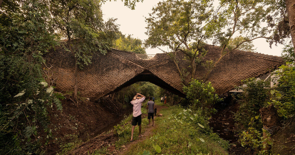 Aerial view of a residential bridge clad in thatch and mud spanning a valley in Karjat, revealing the steel structure beneath the organic surface.