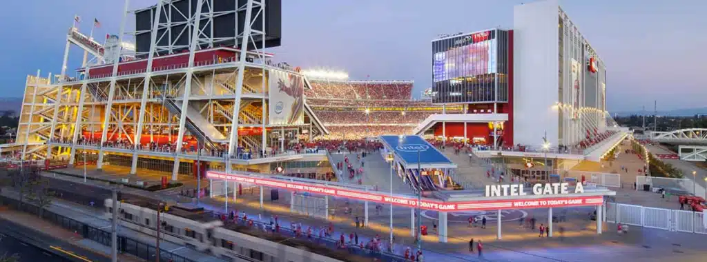 مدخل Intel Gate A في Levi's Stadium عند الغروب، مع إضاءة معمارية مميزة وحشد من الجماهير، وقطار يمر في المقدمة.