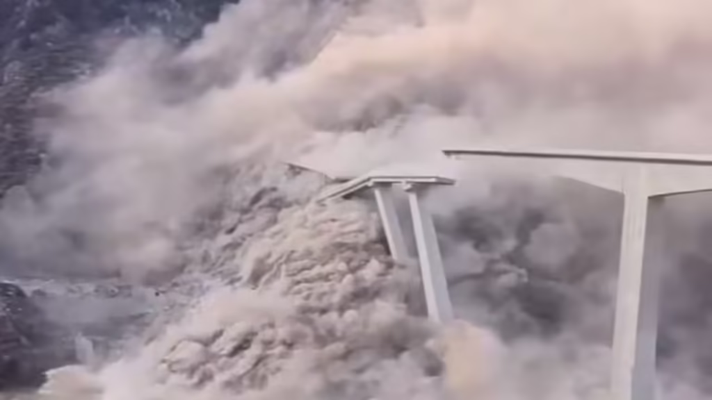 Collapsed bridge showing damaged concrete piers and surrounding mountainous slopes, highlighting the structural impact of the collapse.