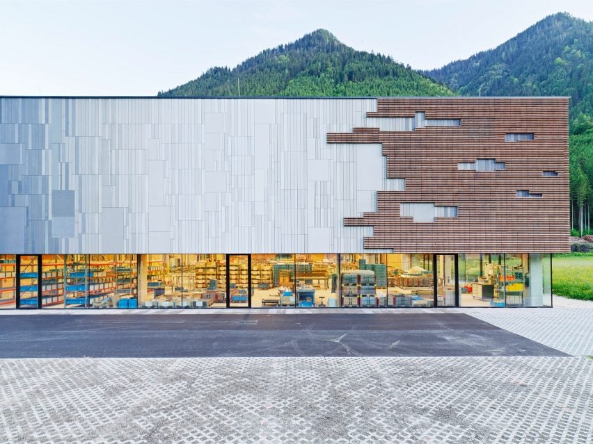 Front view of a modern Austrian production hall with textured metal and timber facade, revealing an illuminated industrial interior against a mountain backdrop.
