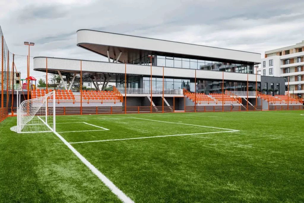 مركز رياضي حديث في ديرلشتي، مُصمم على تلة طبيعية، مع مدرجات برتقالية وأسطح علوية زجاجية تطل على ملعب كرة قدم أخضر.