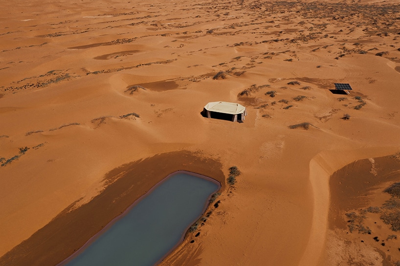 منظر جوي لمشروع ديزرت آرك في صحراء منغوليا الداخلية، حيث ينسلّ الهيكل العضوي بين الكثبان، بجانب بركة ماء ووحدة شمسية، كتذكير بالاندماج مع البيئة.