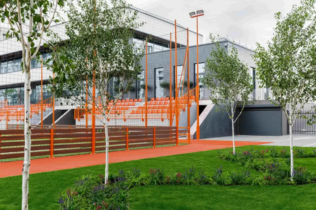 ممر برتقالي مُحيط بمدرجات رياضية وسط حديقة خضراء، مع أشجار بيضاء وأسوار خشبية تربط بين الطبيعة والهندسة المعمارية الحديثة.