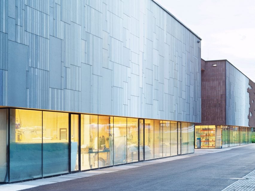 Close up of the hall&rsquo;s metallic facade and glowing glass base, revealing warm interior workspace against a minimalist industrial backdrop