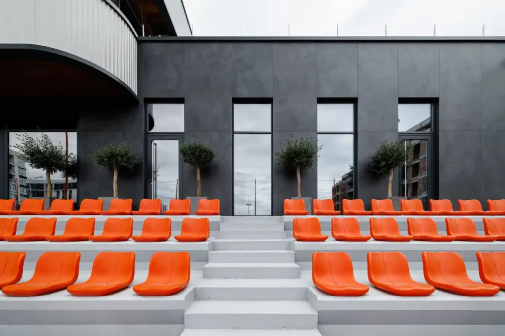 مقاعد برتقالية مُرتبة على درجات خرسانية أمام جدار أسود مُغطى بألواح، مع نوافذ زجاجية وأشجار صغيرة تضيف لمسة طبيعية.