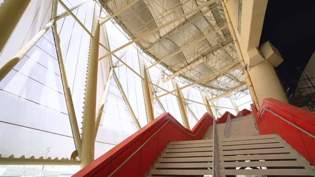 Vibrant red stairs ascend toward a sunlit steel lattice ceiling, guiding the eye upward